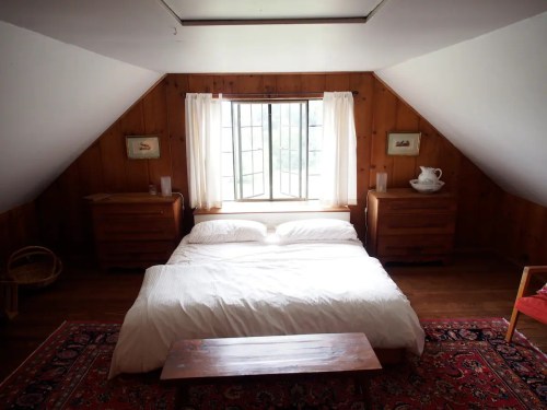 Cozy attic bedroom with a wooden bed, two dressers, a window, and a rug, featuring warm wooden walls and natural light.