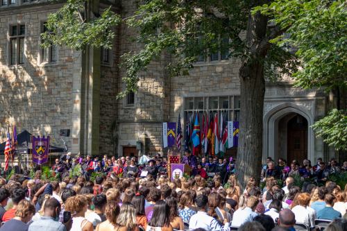 A graduation ceremony outdoors, with graduates seated in front of a stage adorned with flags and university banners.