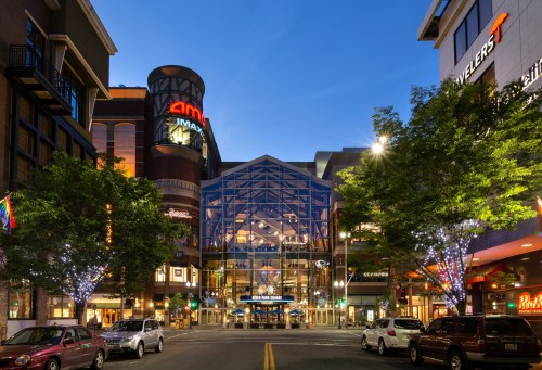 A vibrant shopping center at dusk, featuring a glass entrance, bright lights, and trees adorned with fairy lights.