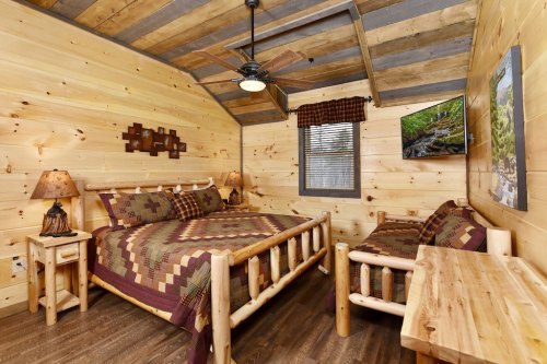 Cozy wooden cabin interior featuring a bed, sofa, and rustic decor with a ceiling fan and wall-mounted TV.
