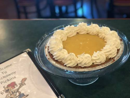 A lemon pie topped with whipped cream sits on a glass cake stand next to a menu on a green table.