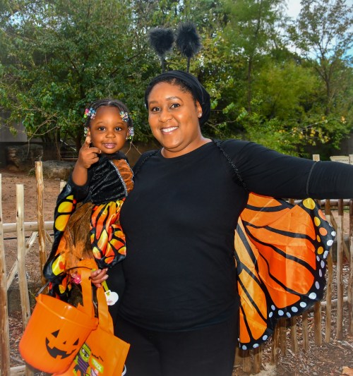 A woman and a young girl in butterfly costumes, smiling and posing outdoors with a Halloween bucket.
