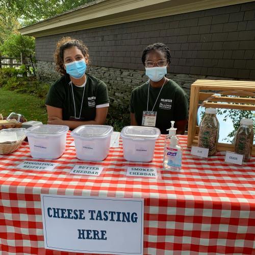 Two volunteers in masks stand behind a table with cheese tasting containers and hand sanitizer, set outdoors.