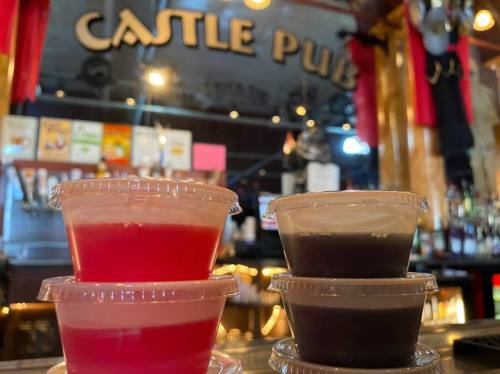 Colorful shot glasses in plastic cups sit on a bar counter with "Castle Pub" sign in the background.