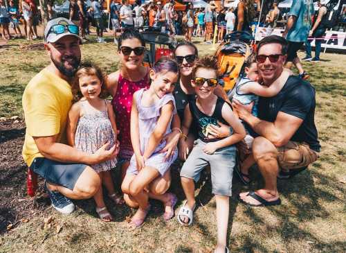 A group of adults and children smiling together at an outdoor event, with strollers in the background.