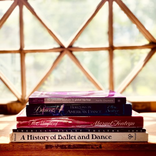 A stack of dance books on a wooden surface, with sunlight streaming through a window in the background.