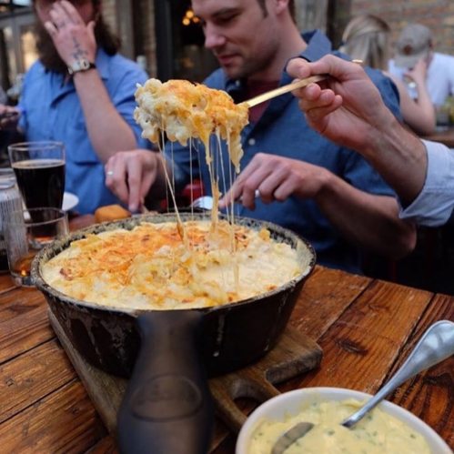 A creamy, cheesy mac and cheese dish being served from a skillet, with drinks and diners in the background.