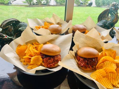Four plates with pulled pork sandwiches and wavy potato chips, set against a green outdoor view.