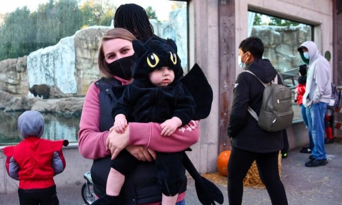 A woman holds a child in a dragon costume at a zoo, with people in the background wearing masks.