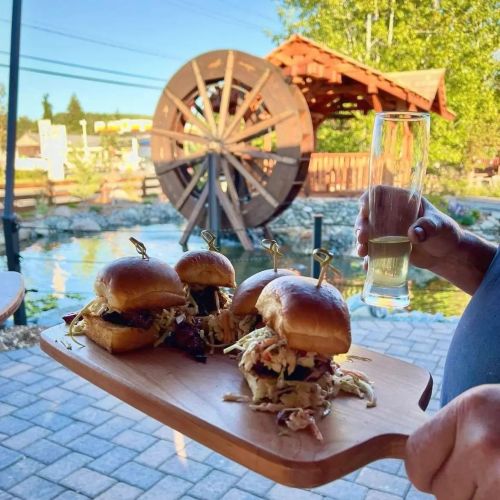 A person holds a wooden platter with three sliders and a drink, with a water wheel and greenery in the background.