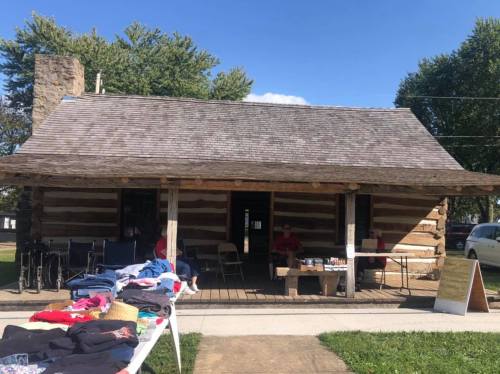 A rustic log cabin with a porch, featuring a clothing sale in front and two people sitting outside.