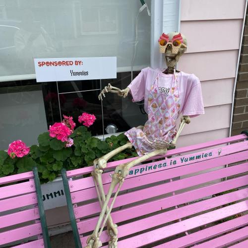 A skeleton in a pink shirt and apron sits on a pink bench, surrounded by flowers, with a sign reading "Sponsored by: Yummies."