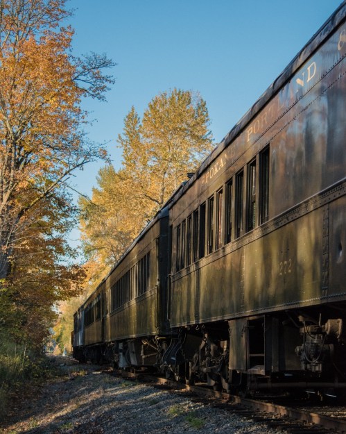 A vintage train car alongside a tree-lined track, surrounded by autumn foliage under a clear blue sky.