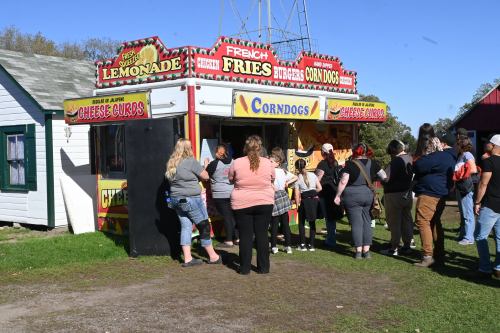 A food stand with a colorful sign offers corndogs, fries, and lemonade, with a line of customers waiting to order.