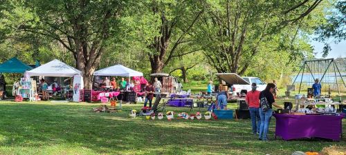 A vibrant outdoor market with tents, vendors, and visitors among trees by a lake on a sunny day.