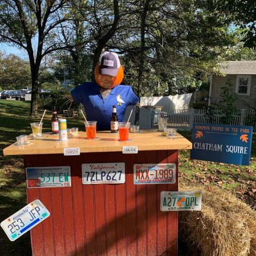 A scarecrow bartender stands at a wooden bar with drinks, surrounded by autumn scenery and a sign for Chatham Square.