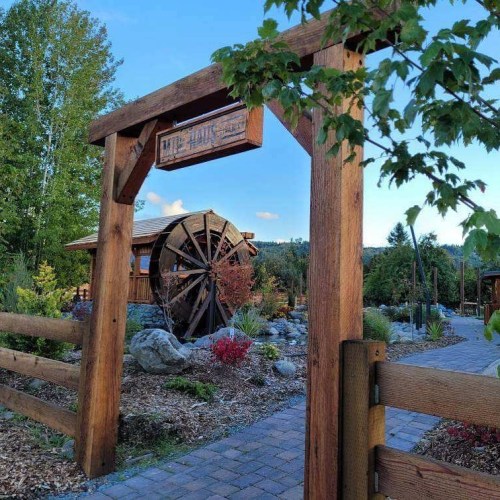 A wooden entrance gate leads to a scenic pathway with a water wheel and lush greenery in the background.