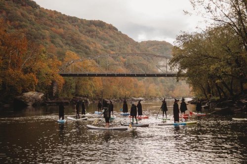 A group of people paddleboarding on a river surrounded by autumn foliage and a bridge in the background.