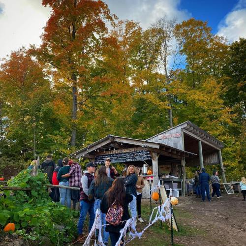 A crowd gathers outside a rustic wooden building surrounded by colorful autumn trees and decorations.