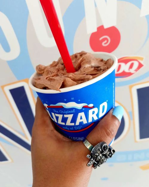 A hand holding a chocolate Blizzard from Dairy Queen with a red straw, against a colorful background.