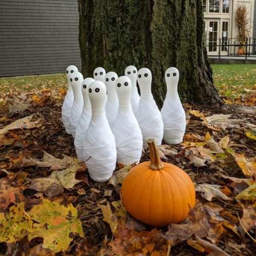 A playful Halloween scene with ghostly bowling pins wrapped in white and a small orange pumpkin among fallen leaves.