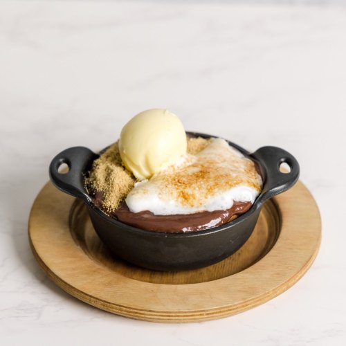 A small cast-iron dish filled with chocolate dessert, topped with meringue and a scoop of vanilla ice cream on a wooden plate.
