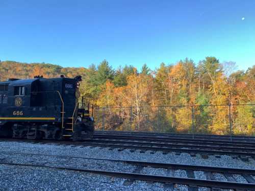 A black train engine parked beside a gravel track, with colorful autumn trees in the background under a clear blue sky.