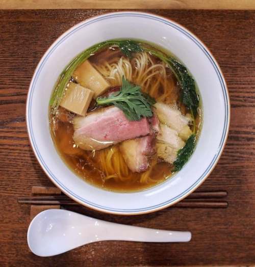 A bowl of ramen with noodles, slices of meat, greens, and bamboo shoots, served on a wooden table with chopsticks.