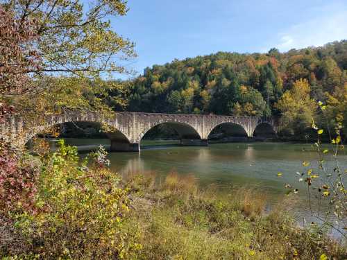 A stone bridge arches over a calm river, surrounded by colorful autumn foliage and trees.