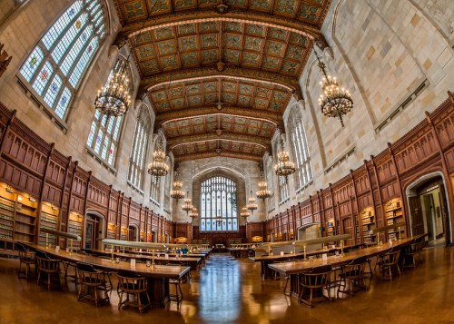 A grand library interior with high ceilings, ornate chandeliers, and wooden bookshelves, featuring long study tables.