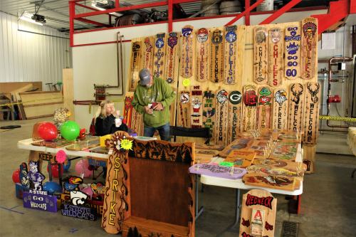 A vendor booth displays colorful wooden signs and crafts, with a man and a child interacting in a workshop setting.