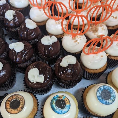 A variety of cupcakes with chocolate and vanilla frosting, some topped with spooky eye decorations and orange pumpkin toppers.