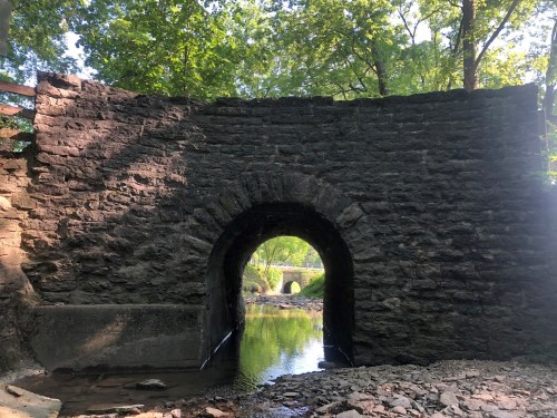 Stone archway leading to a calm stream, surrounded by lush green trees and rocky terrain.