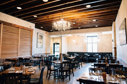 A spacious restaurant interior with wooden beams, chandeliers, and neatly arranged tables and chairs.