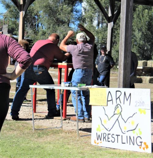 Two men compete in an arm wrestling match under a pavilion, with a sign announcing the event nearby.