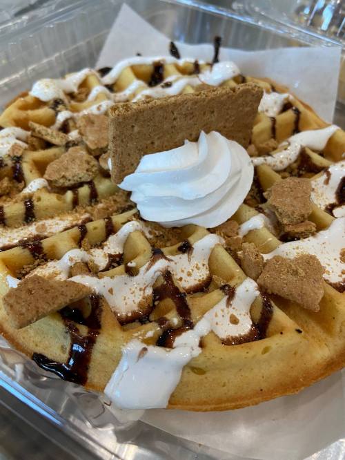 A delicious waffle topped with whipped cream, chocolate drizzle, and crumbled cookies in a clear container.