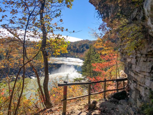 A scenic view of a waterfall surrounded by colorful autumn foliage and rocky cliffs under a clear blue sky.