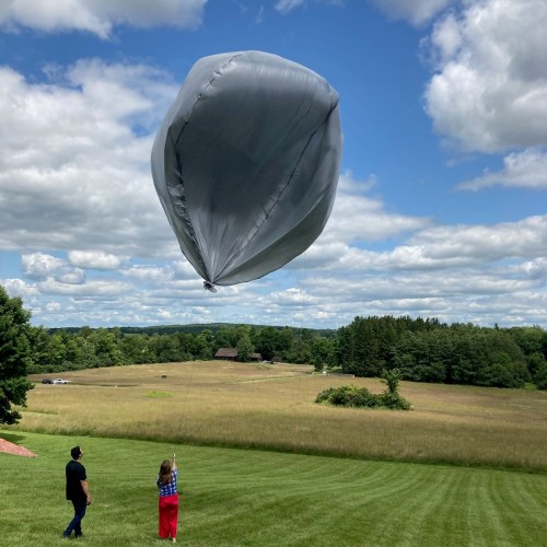 A person holds a tethered gray balloon against a backdrop of clouds and green fields. Another person watches nearby.