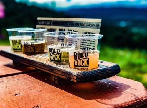 A wooden flight board with three cups of Bold Rock hard cider, set against a scenic mountain backdrop.