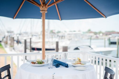 A table set for two under a blue umbrella, featuring plates of food with a marina view in the background.