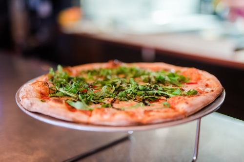 A freshly baked pizza topped with greens on a metal stand, set against a blurred kitchen background.