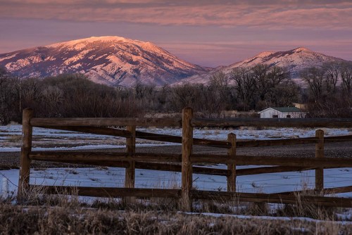 Snow-capped mountains at sunset, framed by a wooden fence and a small house in a serene landscape.