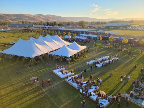 Aerial view of a large outdoor event with tents, tables, and a crowd gathered on a grassy field at sunset.