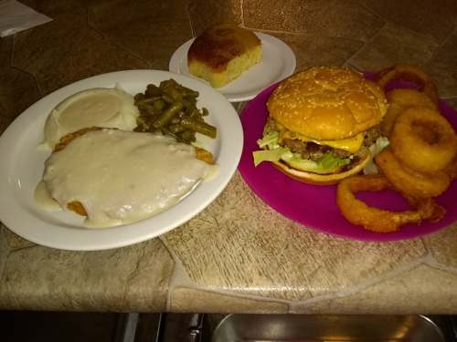 A plate with fried chicken smothered in gravy, mashed potatoes, green beans, and a burger with onion rings and cornbread.