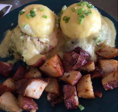 Two eggs Benedict on English muffins with hollandaise sauce, topped with green onions, served with roasted potatoes.