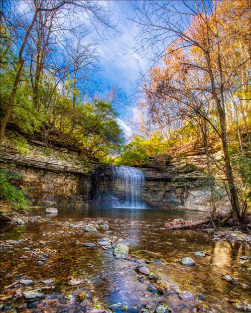 A serene waterfall cascades into a rocky pool, surrounded by lush trees and vibrant autumn foliage under a blue sky.