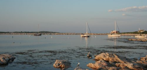 A serene coastal scene with sailboats anchored near rocky shores under a clear sky.