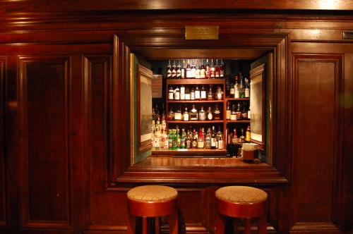 A wooden bar with two stools in front of a shelf filled with various bottles of liquor. Warm, inviting atmosphere.