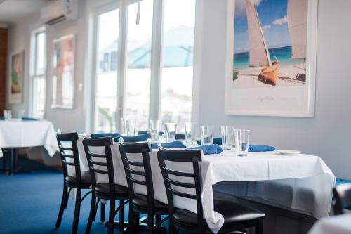 A bright restaurant interior with white tablecloths, empty tables, and beach-themed artwork on the walls.