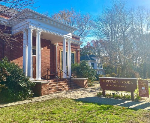 A brick building with white columns, sign reading "Heritage Books at the Old Library," surrounded by greenery.